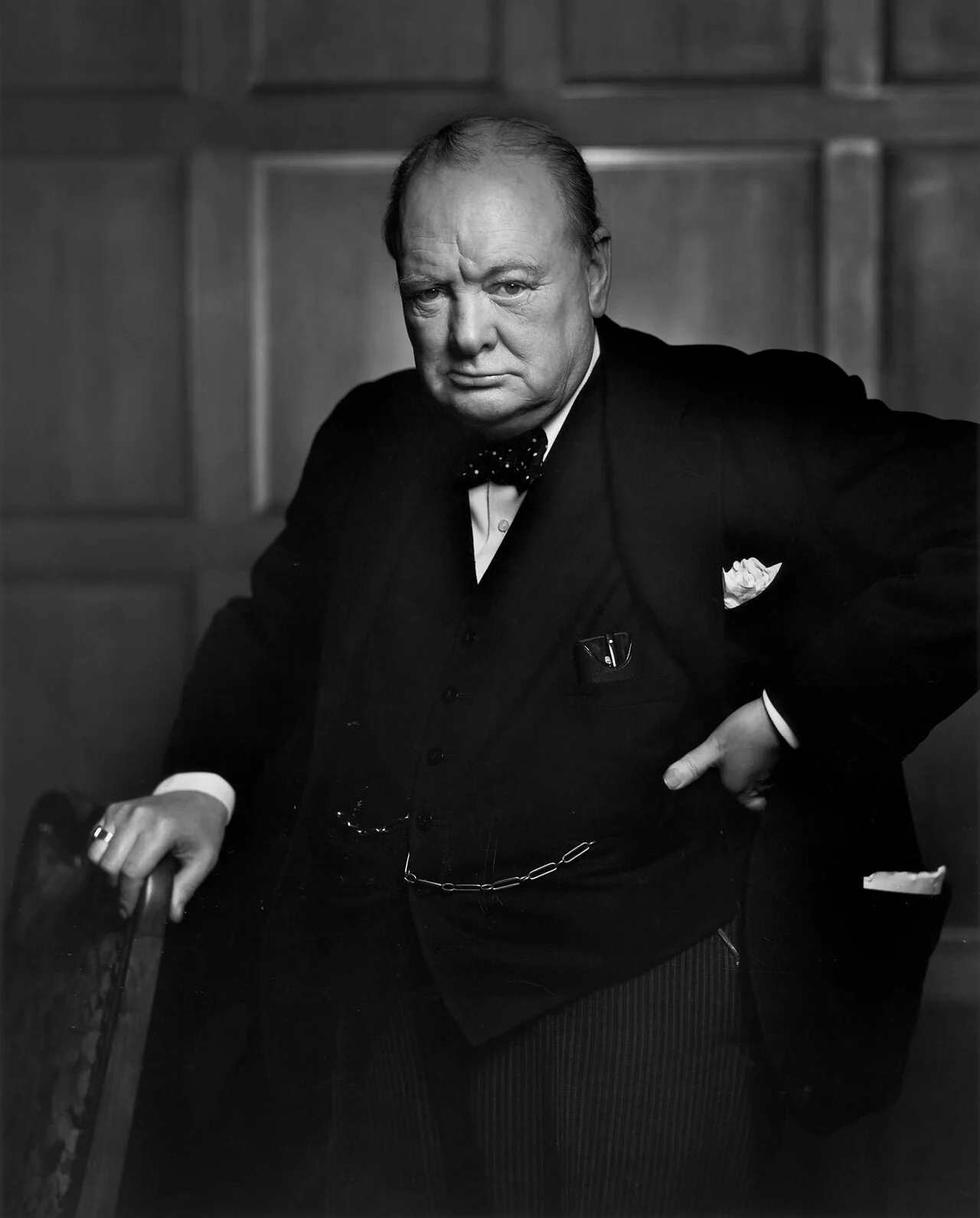 Winston Churchill in a dark suit and spotted bow tie, one hand on a chair and the other on his hip, scowling at the camera.